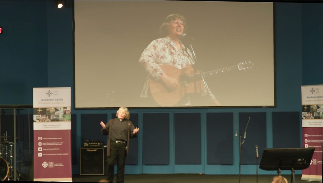 Sarah Jones wearing clericals in front, centre with a visual of her playing guitar above her.